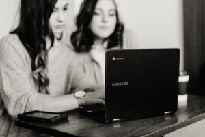 Two young adult women sitting at a desk seemingly working together with one typing on a laptop.
