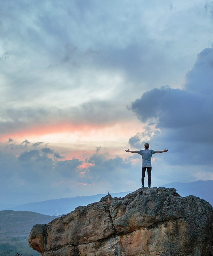 Image of man standing on mountain with outstretched arms and blue skies in front of him.